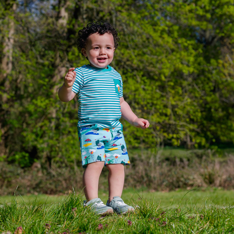 Happy, smiling baby outside, wearing a pair of Ducky Zebra colourful, seaside shorts with a turquoise stripe t-shirt