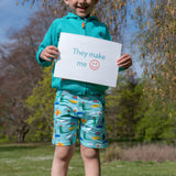 Smiling, happy boy outside holding a sign saying "They make me happy" wearing a pair of Ducky Zebra colourful, seaside print shorts with a turquoise hoody