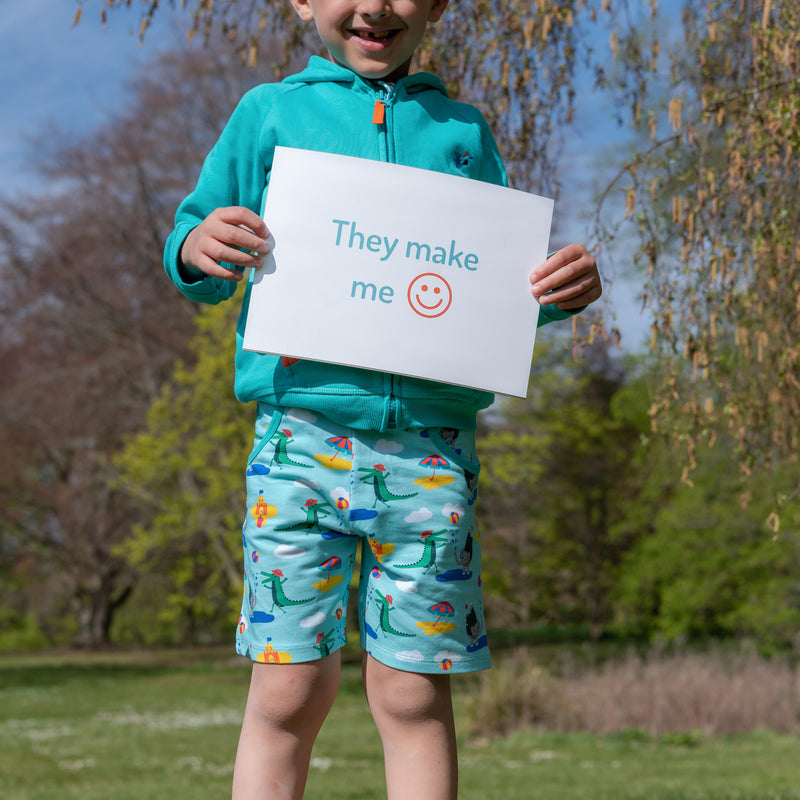 Smiling, happy boy outside holding a sign saying "They make me happy" wearing a pair of Ducky Zebra colourful, seaside print shorts with a turquoise hoody