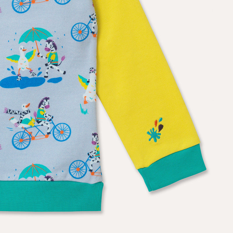 Image of kids raglan sweatshirt with yellow sleeves and repeat print pattern body. The repeat print pattern has images of a duck and zebra splashing in a puddle together, cycling on a bike and running a three legged race. Image focuses on embroidered splash motif on sleeve