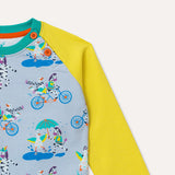 Image of kids raglan sweatshirt with yellow sleeves and repeat print pattern body. The repeat print pattern has images of a duck and zebra splashing in a puddle together, cycling on a bike and running a three legged race. Image focuses on the neckline with two orange buttons to allow easy dressing