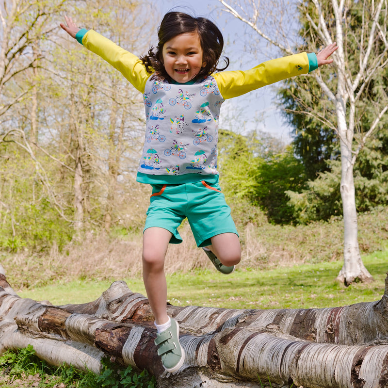 Image of a smiling, happy girl jumping off a tree with her arms spread wide. She is wearing a pair of unisex turquoise shorts and a Ducky Zebra sweatshirt with bright yellow sleeves