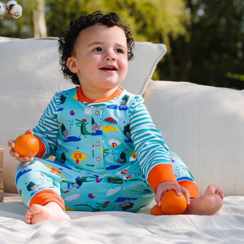 Image of a baby sitting on a blanket outside, wearing a Ducky Zebra colourful sleeveless romper with a turquoise background and repeat print pattern of a happy crocodile and elephant splashing at the seaside. The baby is playing with some oranges