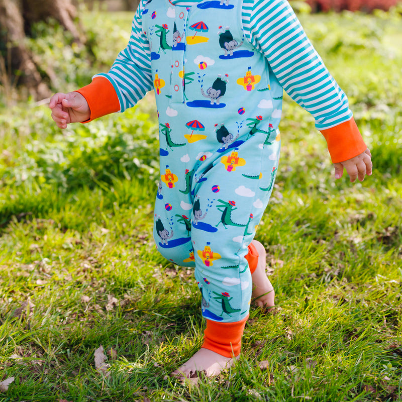 Image of a baby walking in the grass, wearing a Ducky Zebra colourful sleeveless romper with a turquoise background and repeat print pattern of a happy crocodile and elephant splashing at the seaside