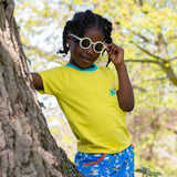 Image of a smiling girl wearing a Ducky Zebra yellow t-shirt and colourful skort, standing by a tree outside