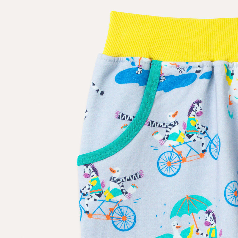 Brightly coloured joggers with a light grey background and repeat print pattern of a duck and zebra splashing in puddles, riding a bike and running a three legged race. Image is zoomed in on the bright yellow elasticated waistband and turquoise pocket trim