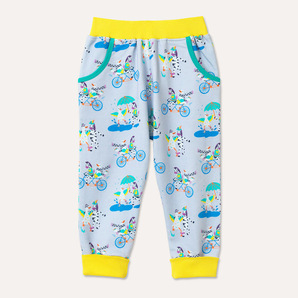 Brightly coloured joggers with a light grey background and repeat print pattern of a duck and zebra splashing in puddles, riding a bike and running a three legged race. Bright yellow elasticated waistband and roll-up cuffs with a turquoise pocket trim