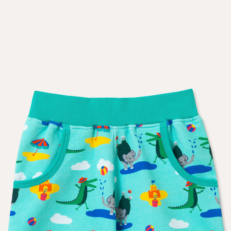 Brightly coloured children's shorts with a turquoise background and repeat print pattern of a smiling crocodile and elephant playing at the seaside. Front shot of the upper half of the shorts, focusing on the two deep pockets and elasticated turquoise waistband