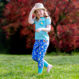 Image of a smiling, happy girl running through the grass with bare feet, wearing a pair of Ducky Zebra colourful joggers and a turquoise stripe t-shirt. The girl is holding onto her straw hat to stop it from flying off