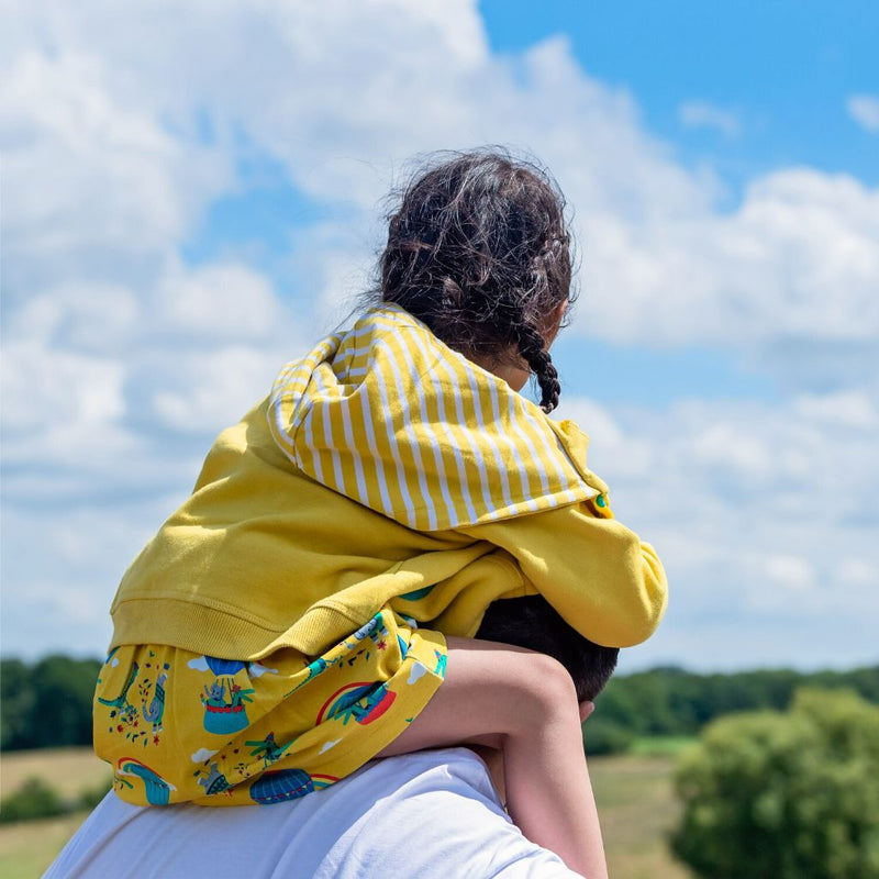 Image of a dad carrying a girl on his shoulders. The girl is wearing a Ducky Zebra dress and yellow hoodie with a white and yellow striped hood lining.