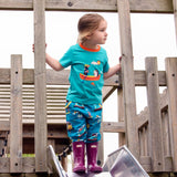 Girl standing at the top of a slide wearing a Ducky Zebra turquoise tshirt and teal joggers with a repeat print pattern of paddleboards and campervans. She's wearing purple wellies. 