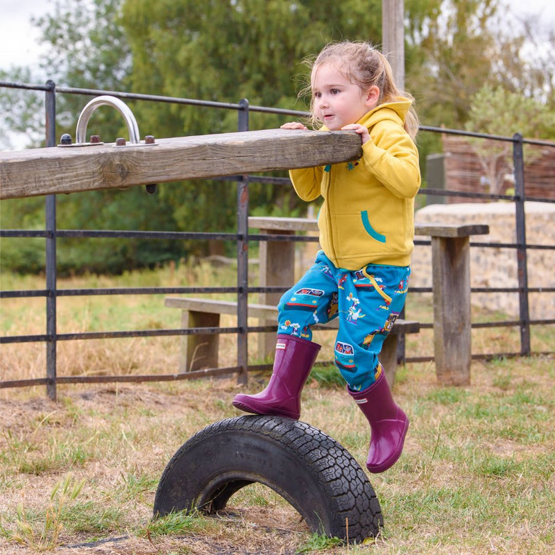 Girl playing on a seesaw, wearing a Ducky Zebra yellow hoodie with colourful, unisex trousers with a campervan and paddleboard repeat print. She's wearing purple wellies.