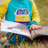 Kid sitting on the grass reading a book wearing a Ducky Zebra unisex jumper. The jumper has an applique of a fox and elephant driving a yellow campervan.
