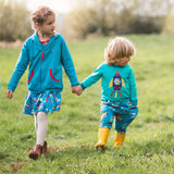 A young brother and sister holding hands outside, both wearing colourful, sustainable Ducky Zebra clothes including a teal hoodie and a turquoise top with a rocket print.