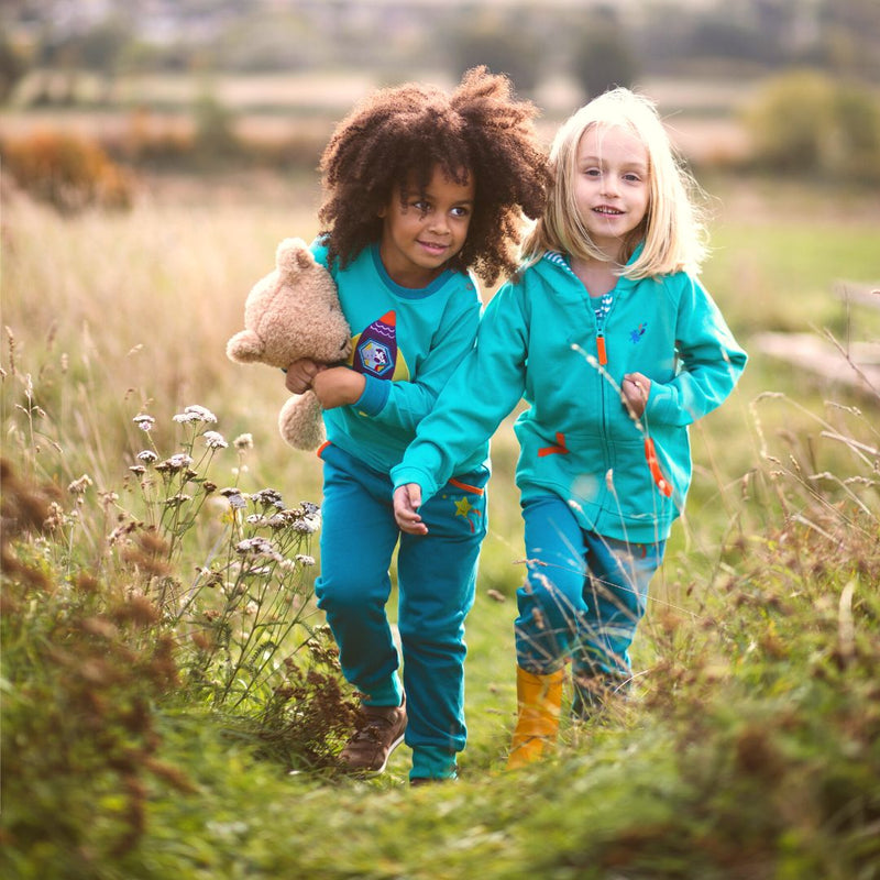 A girl and boy walking together through the long grass, both wearing Ducky Zebra teal joggers and a turquoise top.