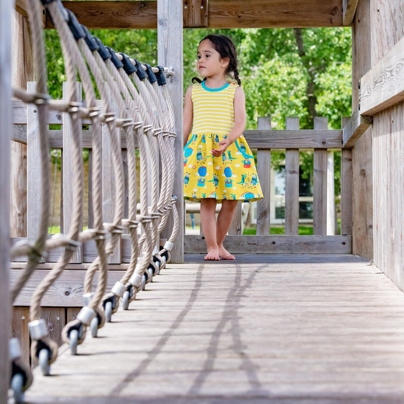 Image of a barefoot girl running across a wooden bridge wearing a Ducky Zebra sleeveless dress. The dress has a yellow and white stripe top half with a printed bottom half. The print has a yellow background with a crocodile and elephant enjoying a hot air balloon ride and playing together in the leaves.