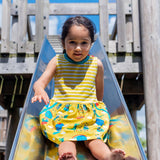 Image of a happy girl on a slide, wearing a Ducky Zebra sleeveless dress. The dress has a yellow and white stripe top half with a printed bottom half. The print has a yellow background with a crocodile and elephant enjoying a hot air balloon ride and playing together in the leaves.