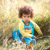Toddler sitting in the long grass wearing a colourful ducky Zebra top and teal joggers.