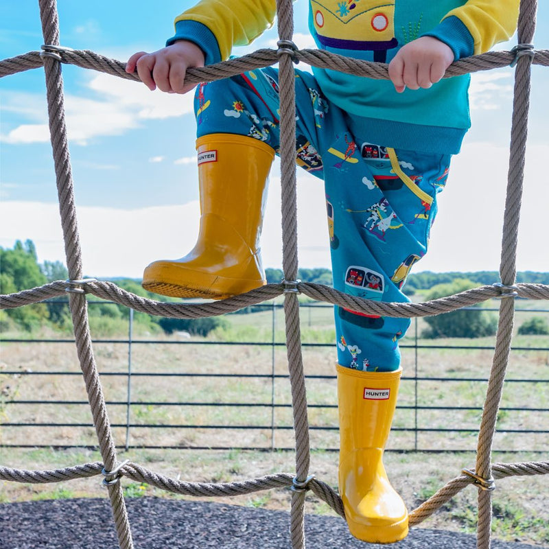Child climbing up a rope net wearing Ducky Zebra teal trousers with a repeat print pattern of campervans and paddleboards. The trousers have a yellow trim around the pocket. The child is wearing yellow wellies. 