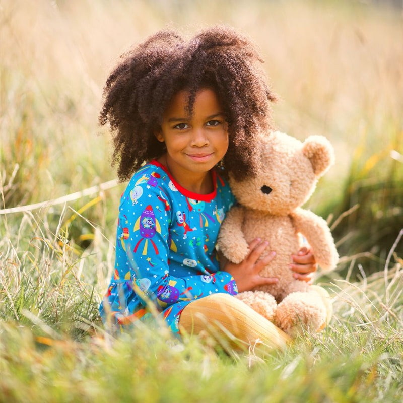 Image of a happy girl sitting in long grass wearing a Ducky Zebra long sleeve skater dress with rockets and stars, cuddling onto a teddy