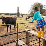 Image of a girl standing on a style by a field of cows. She's wearing a colourful Ducky Zebra skirt and hoody with a pair of wellies 