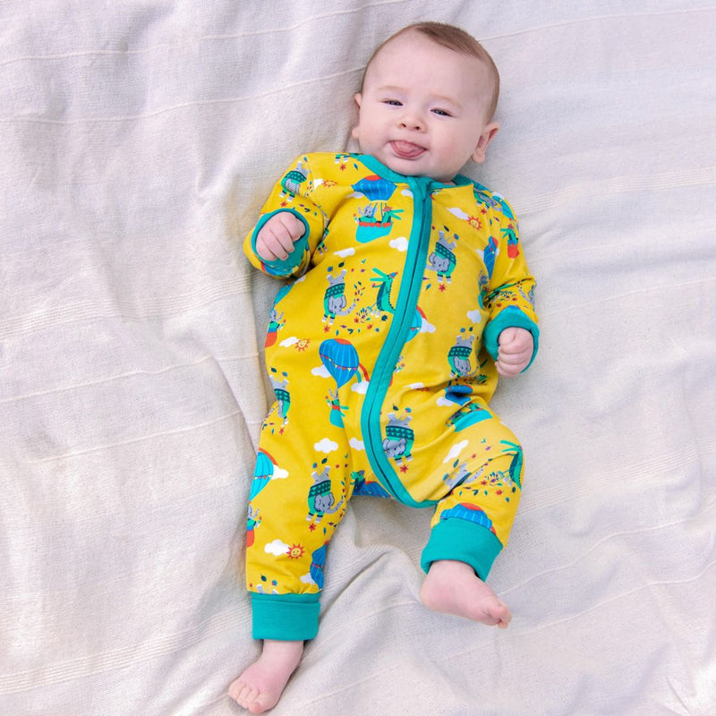 Image of a baby lying on a blanket wearing a Ducky Zebra sleepsuit. The sleepsuit has a yellow background with a repeat print pattern of a crocodile and elephant enjoying a hot air balloon ride and playing in the leaves. The sleepsuit has a contrasting turquoise two-way zip and roll-down cuffs.