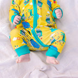 Close-up image of a baby lying on a blanket wearing a Ducky Zebra baby sleepsuit. The sleepsuit has a yellow background with a repeat print pattern of a crocodile and elephant enjoying a hot air balloon ride and playing in the leaves. The sleepsuit has a contrasting turquoise two-way zip and roll-down cuffs.