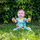 Image of a smiling baby sitting in the grass, wearing a Ducky Zebra baby romper, with a light green background and repeat print pattern of a dog and hedgehog rowing and carrying their boat. The romper has a contrasting yellow 2-way zip and cuffs