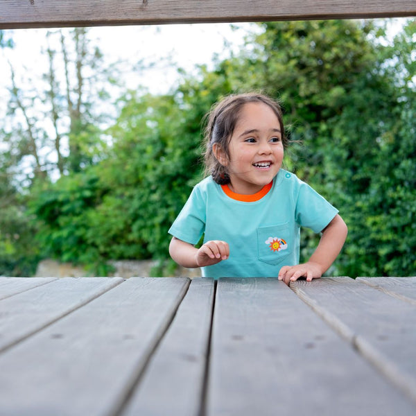 Image of a smiling girl at the top of a climbing frame wearing a light green Ducky Zebra t-shirt with a contrasting orange trim on the neck. The t-shirt has a front pocket on the right hand side with a fun rainbow print. 