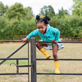 Image of a child climbing over a fence wearing a Ducky Zebra turquoise hoodie and green skort. The girl is also wearing bright yellow wellies