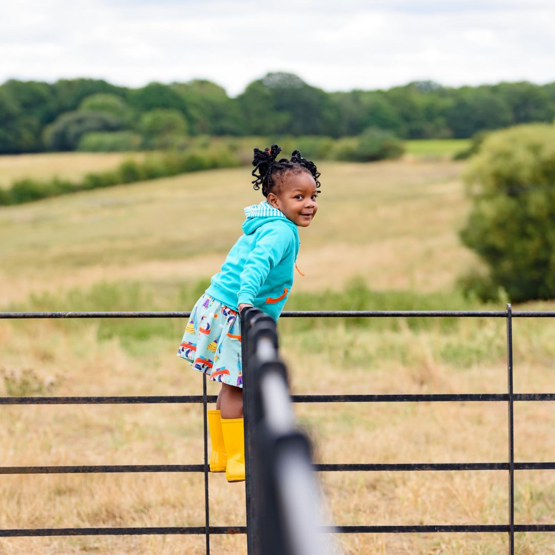 Image of a smiling girl standing on a fence wearing a colourful skort with a canoeing dog and hedgehog print with a turquoise hoodie and bright yellow wellies 