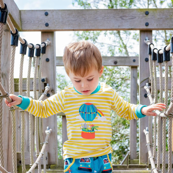 Image of a boy balancing on a wooden bridge, wearing a Ducky Zebra long-sleeve t-shirt. The t-shirt has yellow and white stripes with a print of a crocodile and elephant enjoying a hot air balloon ride together. There are two orange poppers on one of the shoulders. 