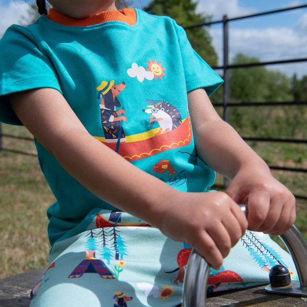 Image of a child wearing a turquoise Ducky Zebra t-shirt with a contrasting orange trim and a print of a hedgehog and sausage dog rowing a boat on the front.